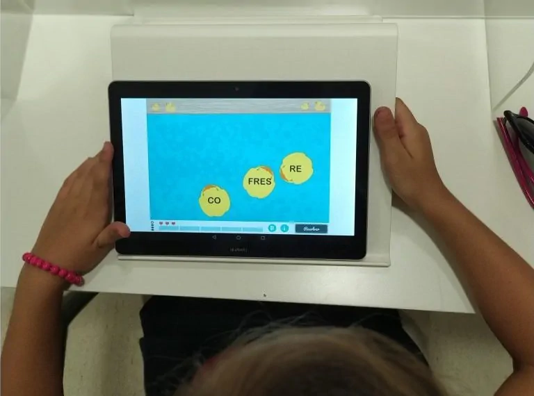 Profissional usando um tablet em ambiente escolar; tela azul com letras e círculos amarelos, pulseira rosa e canetas.