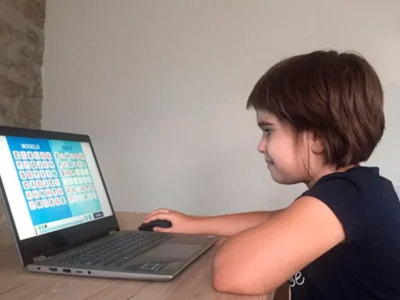 Menina sentada diante de um portátil, interagindo com uma atividade educativa de letras na tela, em um ambiente interno.