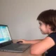 Menina sentada diante de um portátil, interagindo com uma atividade educativa de letras na tela, em um ambiente interno.