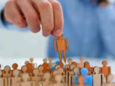 Mão segurando uma figura laranja sobre um grupo de figuras bege e azuis, todas em estilo de silhuetas simples.