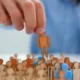 Mão segurando uma figura laranja sobre um grupo de figuras bege e azuis, todas em estilo de silhuetas simples.