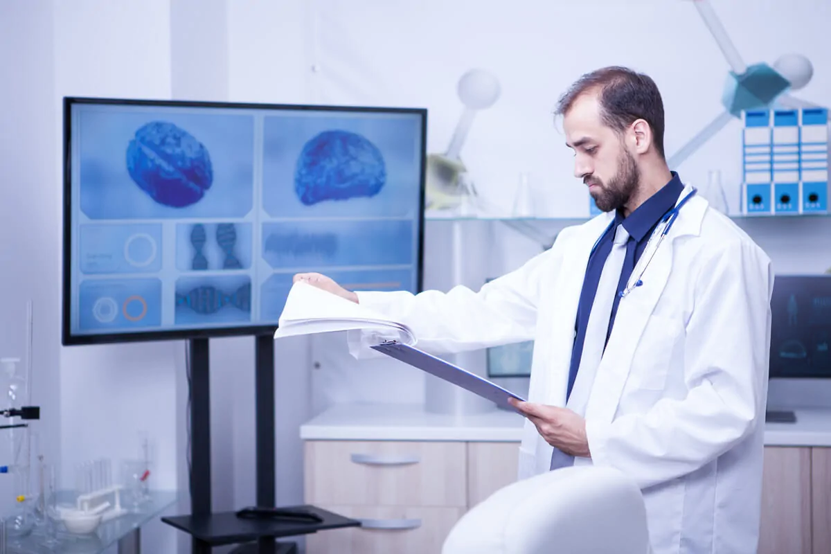 Investigador com bata revisa documentos em laboratório, diante de uma tela com dados para demência frontotemporal.