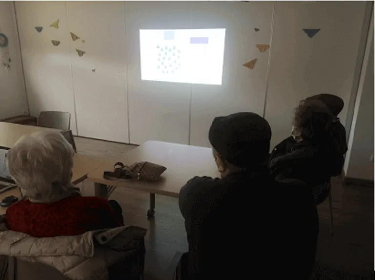Grupo de pessoas idosas sentadas em uma sala, olhando uma apresentação projetada na parede com gráficos e decorações geométricas.