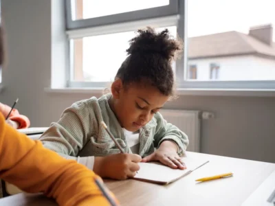 Menina concentrada escrevendo em um caderno em uma sala de aula; cabelo cacheado, camiseta branca e jaqueta verde, janela com luz natural.