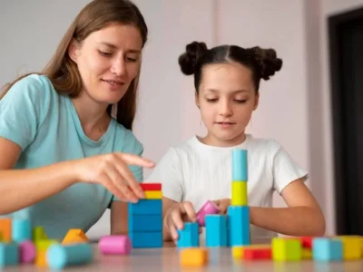 Mulher e menina brincando com blocos coloridos em uma mesa; imagem educativa que ilustra a interação e o desenvolvimento infantil.