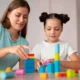 Mulher e menina brincando com blocos coloridos em uma mesa; imagem educativa que ilustra a interação e o desenvolvimento infantil.