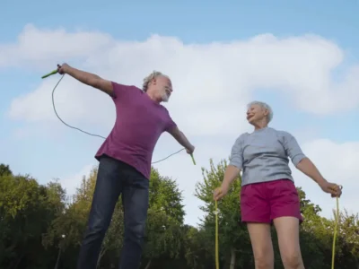 Dois idosos em um parque praticando exercícios; um homem pula corda enquanto uma mulher o observa, sob céu claro.