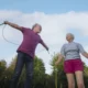 Dois idosos em um parque praticando exercícios; um homem pula corda enquanto uma mulher o observa, sob céu claro.