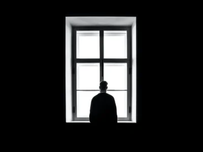 Figura humana de costas para uma janela grande iluminada; quarto escuro e luz suave que inspira contemplação.