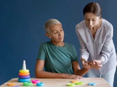 Duas mulheres em um ambiente interno, uma está sentada observando a outra que explica junto a um brinquedo empilhável e peças geométricas sobre uma mesa azul.