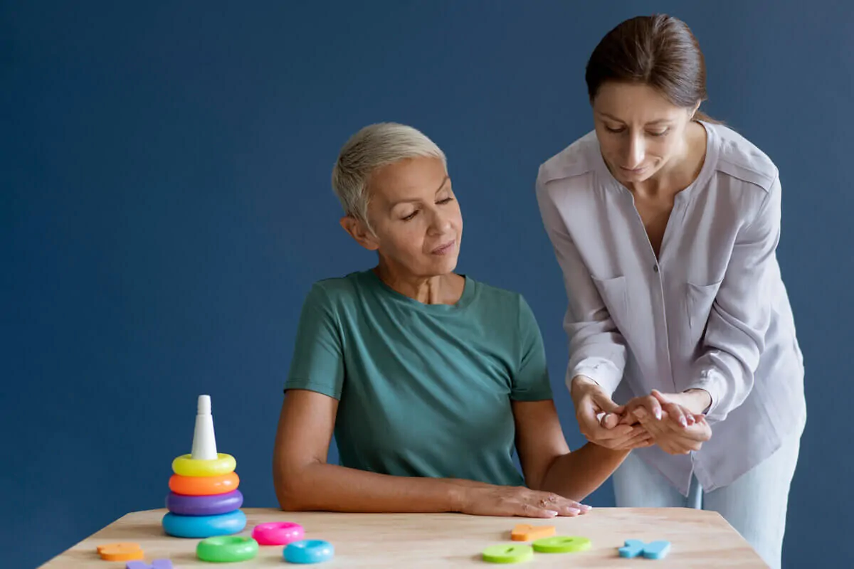 Duas mulheres em um ambiente interno; uma está sentada observando a outra que explica junto a um brinquedo empilhável e peças geométricas sobre uma mesa azul.