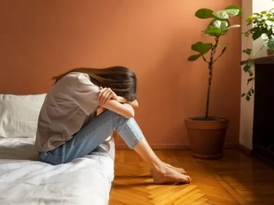 Mulher sentada na cama, cabeça entre os joelhos e olhar reflexivo, em um ambiente quente com uma planta e luz suave.