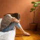Mulher sentada na cama, cabeça entre os joelhos e olhar reflexivo, em um ambiente quente com uma planta e luz suave.