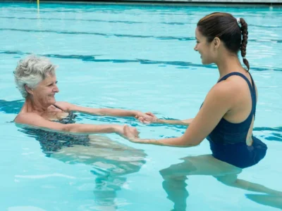 Duas mulheres em uma piscina: uma mulher mais idosa recebe apoio da treinadora para exercícios na água, e ambas sorriem.