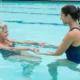 Duas mulheres em uma piscina: uma mulher mais idosa recebe apoio da treinadora para exercícios na água, e ambas sorriem.