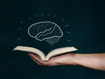 Mão segurando um livro aberto com um cérebro desenhado flutuando sobre as páginas e linhas de ideias que emanam.