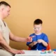 Profissional da saúde interage com uma criança durante uma sessão de jogo terapêutico em uma mesa cercada de brinquedos.