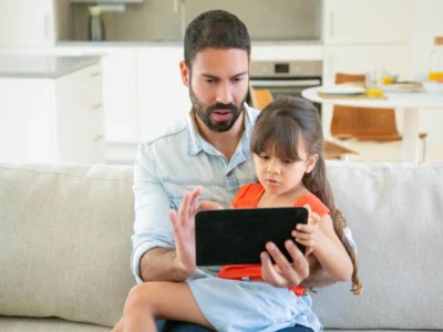 Um homem barbudo e uma menina sentados em um sofá observam um tablet, com uma cozinha iluminada ao fundo.