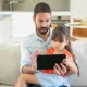 Um homem barbudo e uma menina sentados em um sofá observam um tablet, com uma cozinha iluminada ao fundo.