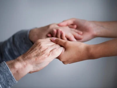 Mãos entrelaçadas em primeiro plano, uma pessoa idosa e outra mais jovem, com fundo desfocado.
