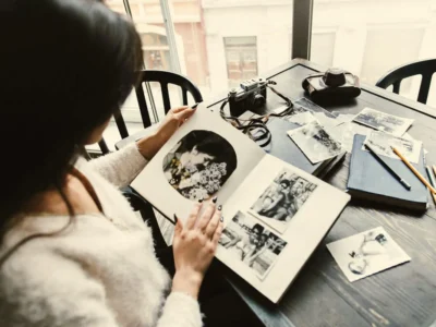 Pessoa sentada à mesa olhando um álbum de fotos em preto e branco, cercada de cadernos, fotos soltas e uma câmera.