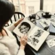 Pessoa sentada à mesa olhando um álbum de fotos em preto e branco, cercada de cadernos, fotos soltas e uma câmera.
