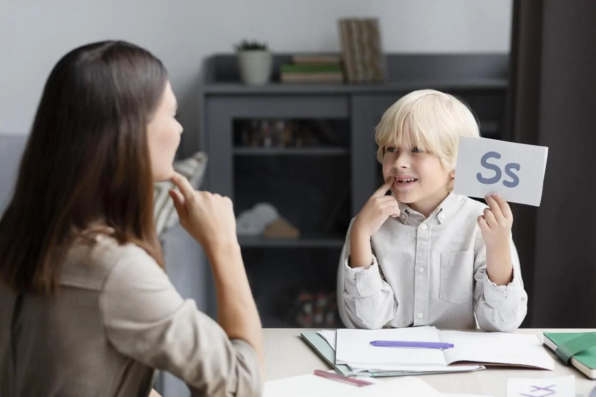 Menino com cartão com a letra S em uma mesa, sob a supervisão de uma profissional; intervenção educativa com a NeuronUP em um tablet.