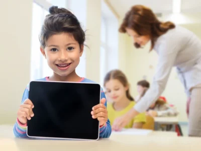 Menina sorrindo segura um tablet em uma sala de aula iluminada, com uma professora e outras meninas ao fundo, durante a estimulação cognitiva.