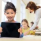 Menina sorrindo segura um tablet em uma sala de aula iluminada, com uma professora e outras meninas ao fundo, durante a estimulação cognitiva.
