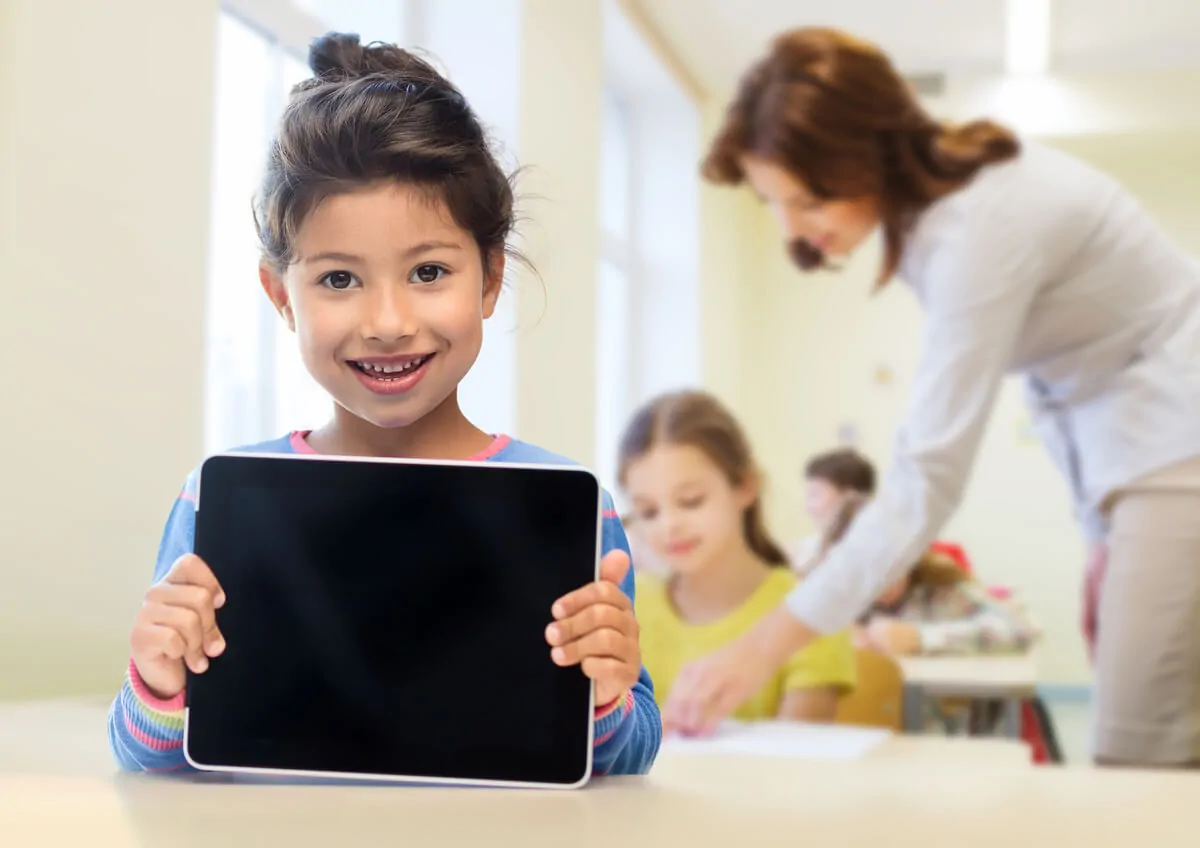 Uma menina sorridente segura um tablet em uma sala de aula iluminada, com uma professora e outras meninas ao fundo, durante a estimulação cognitiva.