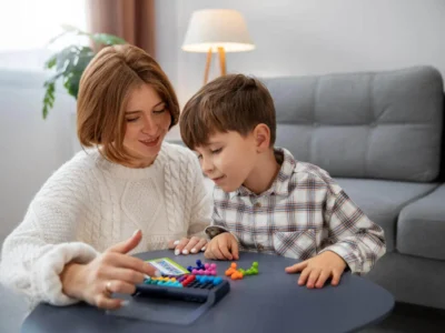 Uma mulher e uma criança interagem durante uma atividade educativa à mesa, em um ambiente doméstico com peças coloridas.