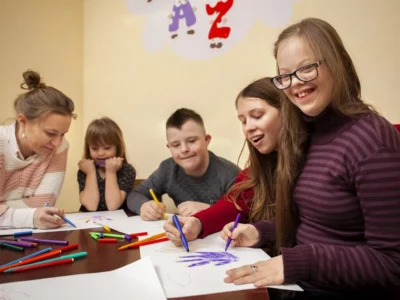 Grupo de três crianças e dois adultos participando de atividades manuais ao redor de uma mesa em um ambiente escolar inclusivo.