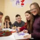 Grupo de três crianças e dois adultos participando de atividades manuais ao redor de uma mesa em um ambiente escolar inclusivo.