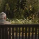 Homem idoso sentado em um banco de madeira em um parque, visto de costas, com ambiente natural e luz suave.