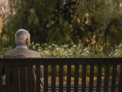 Homem idoso sentado em um banco de madeira em um parque, visto de costas, com ambiente natural e iluminação suave.
