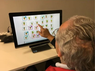Pessoa idosa sentada em frente a um monitor; na tela aparecem ícones de vasos de plantas e a pessoa aponta um deles com o dedo.