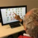 Pessoa idosa sentada em frente a um monitor; na tela aparecem ícones de vasos de plantas e a pessoa aponta um deles com o dedo.