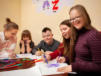 Grupo diverso de alunos e uma docente participam de uma atividade de desenho com marcadores coloridos em uma sala de aula inclusiva.