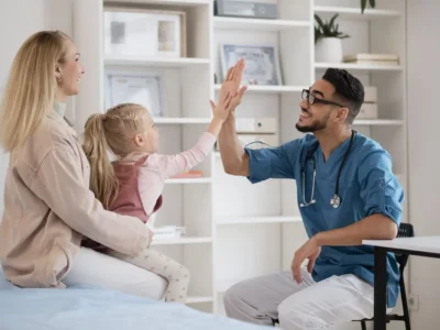Médico em consulta pediátrica sorri e dá um high five a uma menina, enquanto a mãe observa em um ambiente clínico.