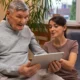 Homem mais velho e mulher jovem sentados juntos; ele segura um tablet, ela lhe explica; fundo com luz natural e plantas.