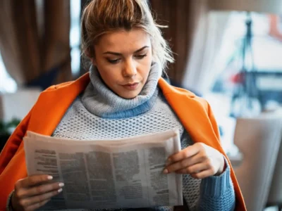Mulher com casaco laranja sentada em um ambiente acolhedor lendo um jornal, com iluminação suave.