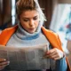Mulher com casaco laranja sentada em um ambiente acolhedor lendo um jornal, com iluminação suave.