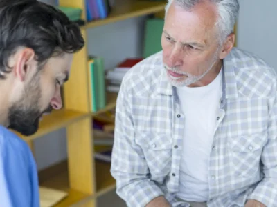 Fonoaudiólogo e um homem adulto em consulta clínica; fundo com estantes e arquivos, durante uma sessão de afasia.