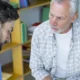 Fonoaudiólogo e um homem adulto em consulta clínica; fundo com estantes e arquivos, durante uma sessão de afasia.