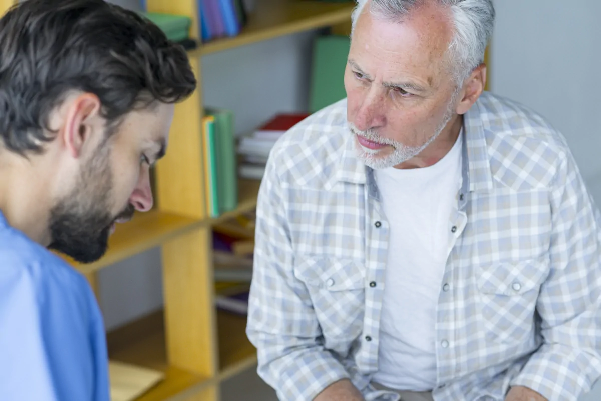 Fonoaudiólogo e um homem adulto em consulta clínica; fundo com prateleiras e arquivos, durante uma sessão de afasia.