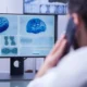 Monitor com gráficos cerebrais e hélices de DNA em azul, rodeado de telas com dados, e uma pessoa de costas falando ao telefone.