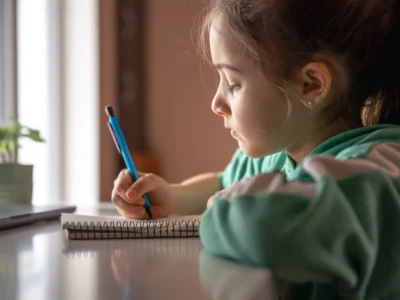 Menina pequena sentada frente a uma mesa, concentrada enquanto escreve em um caderno com lápis azul; veste camiseta verde e há uma planta junto à janela.