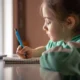 Menina pequena sentada frente a uma mesa, concentrada enquanto escreve em um caderno com lápis azul; veste camiseta verde e há uma planta junto à janela.