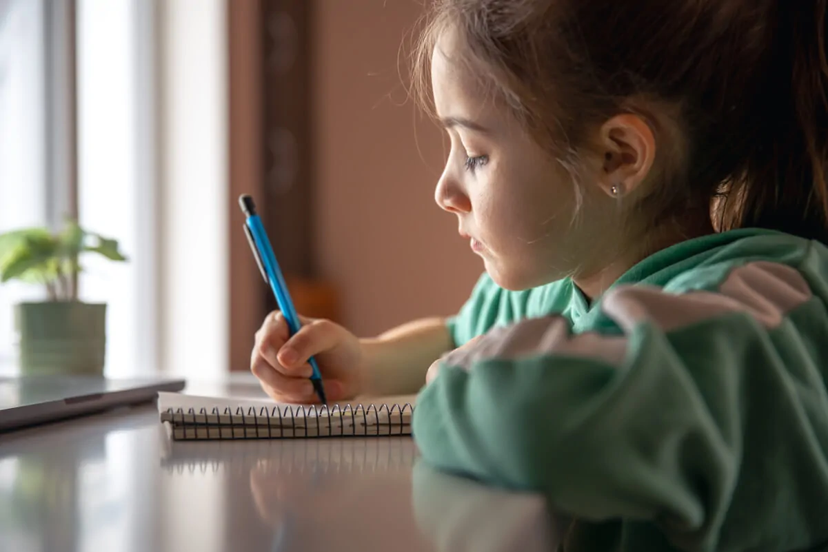 Menina pequena sentada à frente de uma mesa, concentrada enquanto escreve em um caderno com lápis azul; veste camiseta verde e há uma planta ao lado da janela.
