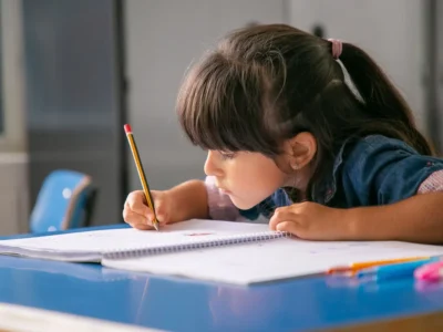 Menina concentrada escrevendo em um caderno com um lápis amarelo, rodeada de canetas coloridas em um ambiente escolar.